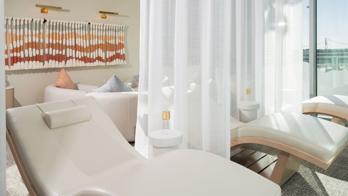 The image shows a serene spa room with lounge chairs, soft pillows, and sheer curtains, suggesting a relaxing environment.