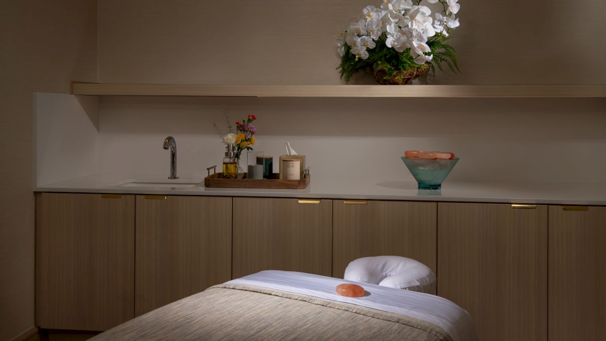 The image shows a serene spa room with a massage table, towels, candles, and decorative flowers on a shelf, creating a calming ambiance.