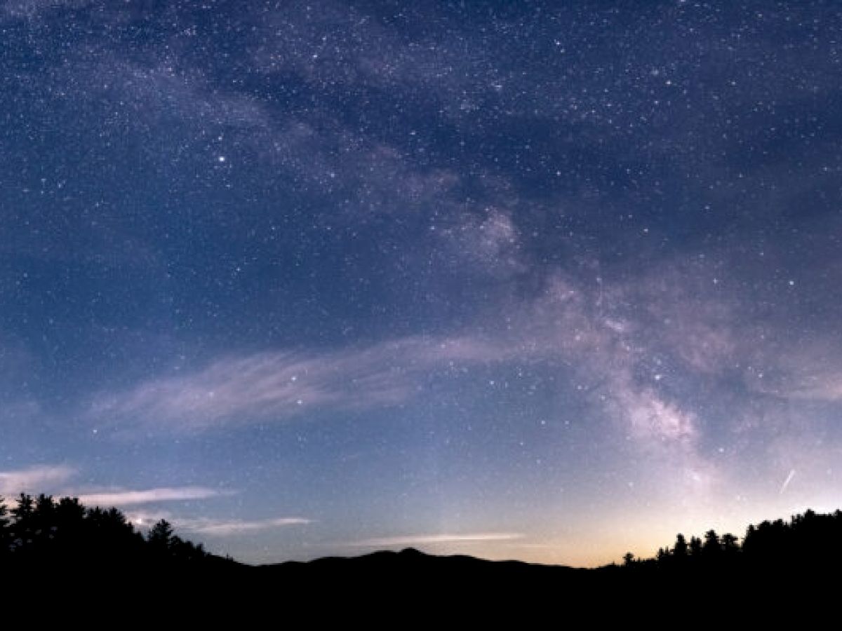 A starry night sky with the Milky Way visible over a silhouetted landscape with trees along the horizon and a gentle glow on the right.