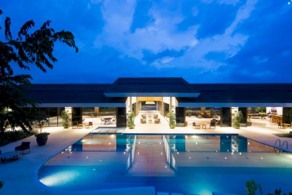 The image shows a luxurious building with an illuminated, large outdoor swimming pool under a twilight sky, surrounded by trees and modern seating areas.