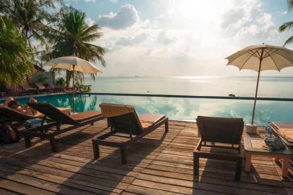 A serene poolside scene with lounge chairs, umbrellas, and palm trees overlooking a calm water body under a partly cloudy sky with sunlight.