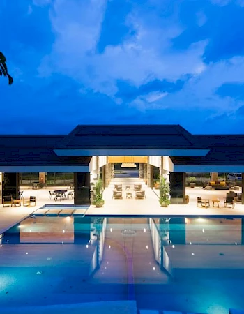 A luxurious resort with a large swimming pool, lounge chairs, and an open-air building in the background, set against a twilight sky.