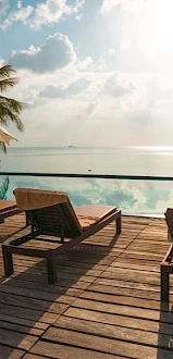 The image shows a serene poolside setting with lounge chairs, umbrellas, and a view of a calm sea under a partly cloudy sky.