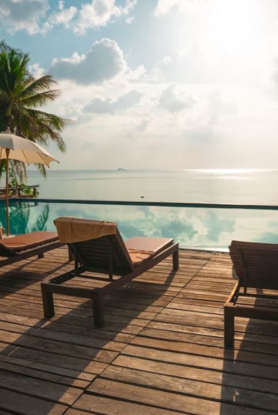 A serene poolside scene with lounge chairs, umbrellas, and palm trees, overlooking a calm body of water under a partly cloudy sky.