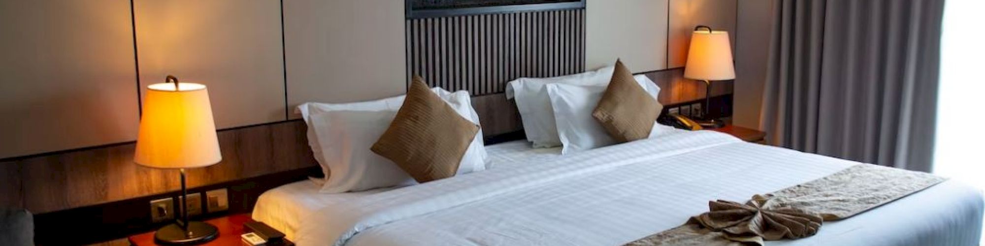 A cozy hotel room with a large bed, bedside tables with lamps, a wall-mounted fireplace, curtains, and decorative pillows and blankets.