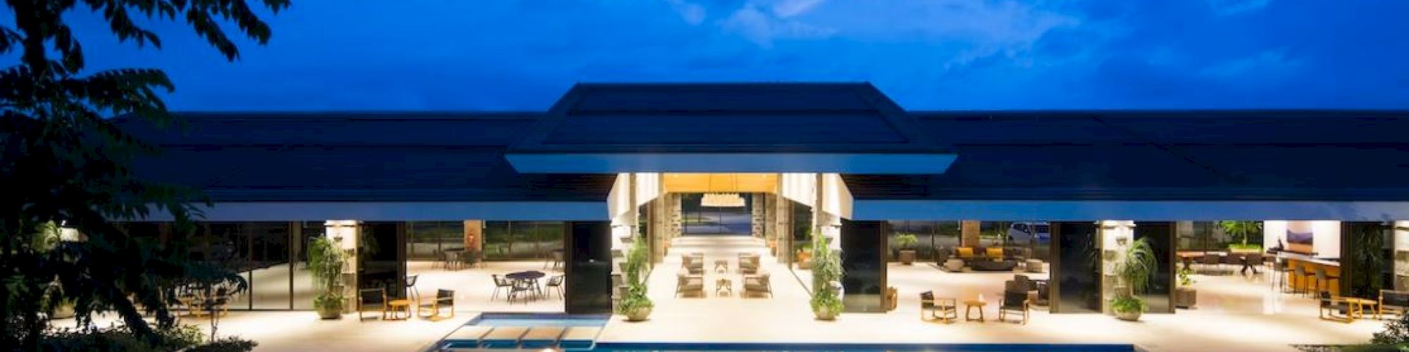 A luxurious swimming pool area is illuminated at dusk, with a modern building featuring open spaces and a serene sky in the background.
