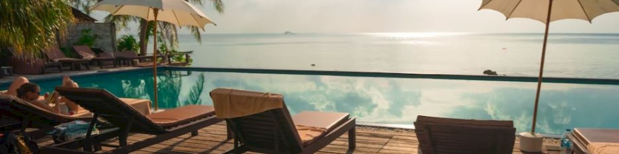 A serene poolside setting with lounge chairs, a table, and umbrellas under a partly cloudy sky, surrounded by palm trees with a view of the water.