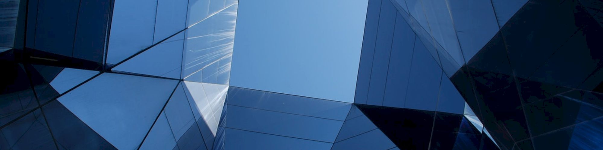 The image shows a view looking up through a geometric arrangement of reflective glass panels with a glimpse of the blue sky in the center.