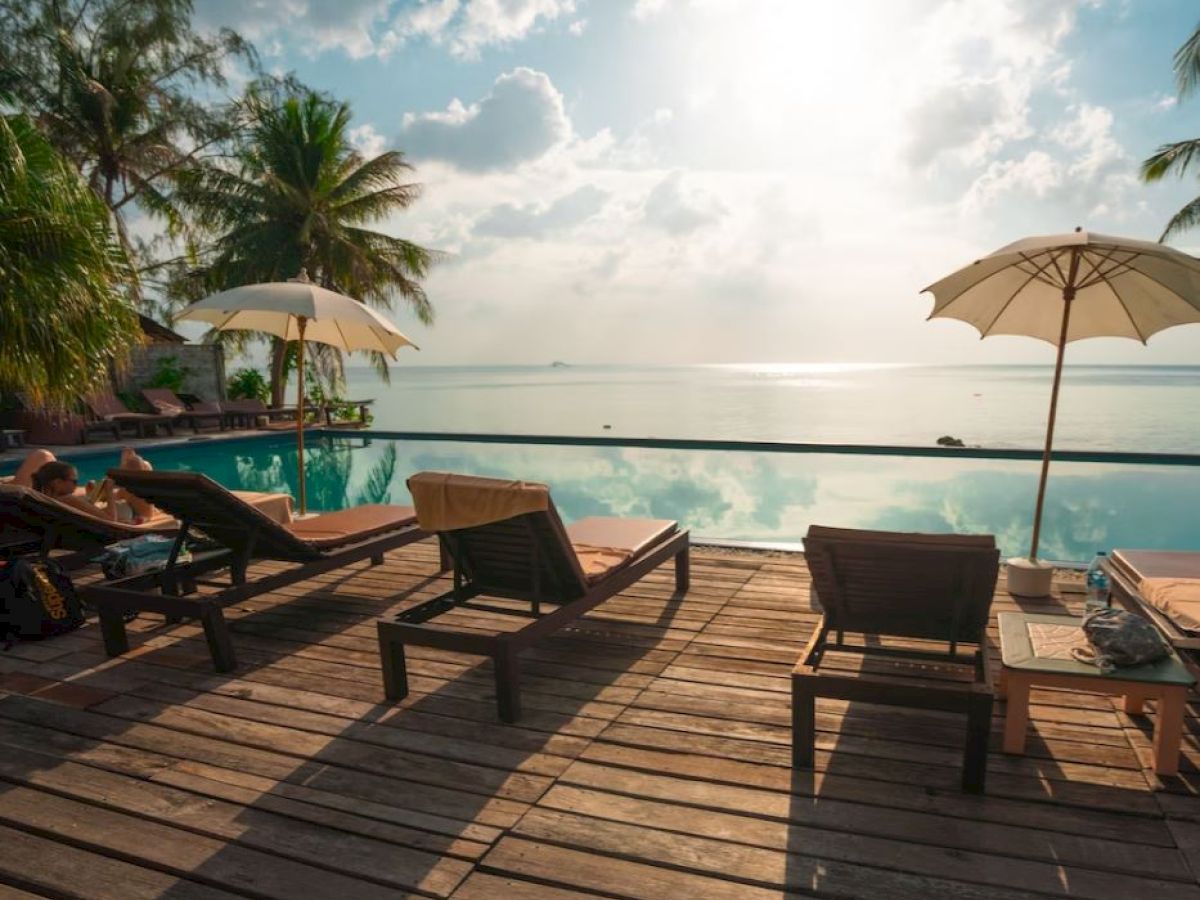 A serene poolside setting featuring lounge chairs, umbrellas, and a view of the ocean under a partly cloudy sky.