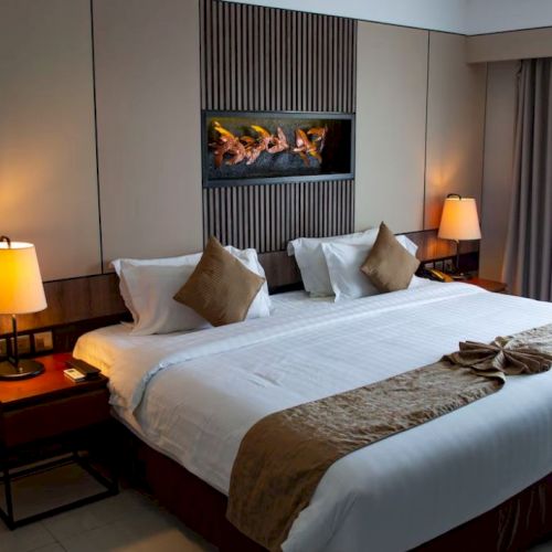 This image shows a hotel room with a large bed, two bedside tables with lamps, a wall-mounted fireplace, and a window with curtains.