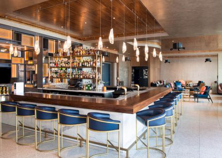 This image shows a modern bar with a wooden counter, blue barstools, hanging lights, and a well-stocked bar. A cozy seating area is in the background.