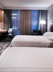 The image shows a modern hotel room with two double beds, a wall-mounted TV, a desk with a chair, lamps, and large windows with curtains.
