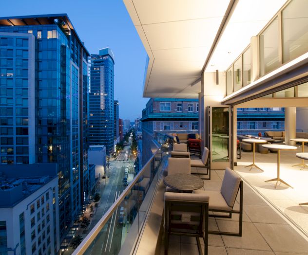 A rooftop terrace with modern furniture is shown in a cityscape during dusk, overlooking tall buildings and a street below, with city lights illuminating.