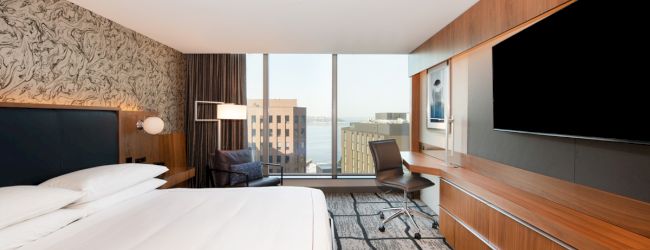 A modern hotel room features a bed, desk, chair, large TV, and a window with a city view, decorated with contemporary furnishings and patterns.