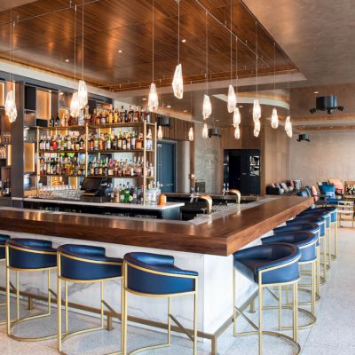 The image shows a modern bar with a wooden counter, high chairs, and various bottles displayed behind the bar. There are elegant lights above.