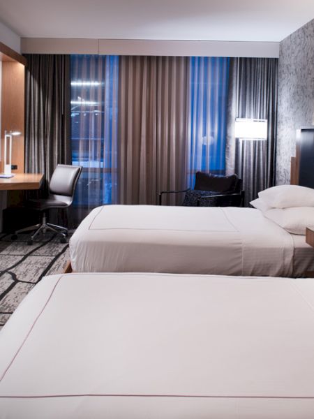 A modern hotel room features two neatly made beds, a flat-screen TV, a desk with a chair, and contemporary decor with a patterned accent wall.
