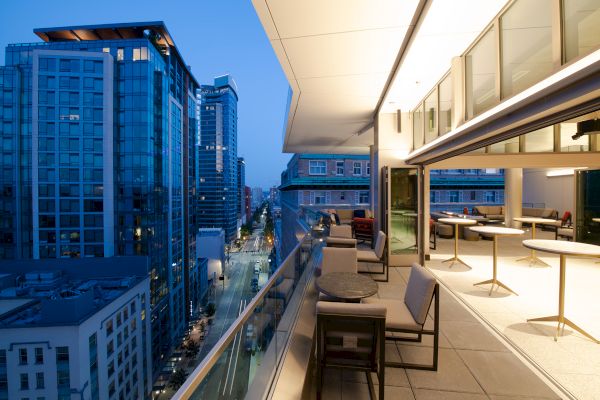 A modern rooftop patio with tables, chairs, and outdoor seating, overlooking a city street with tall buildings and lights during the early evening.