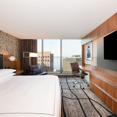 A modern hotel room featuring a large bed, wall-mounted TV, desk, chair, and a window with an urban view, standing out in the scene.