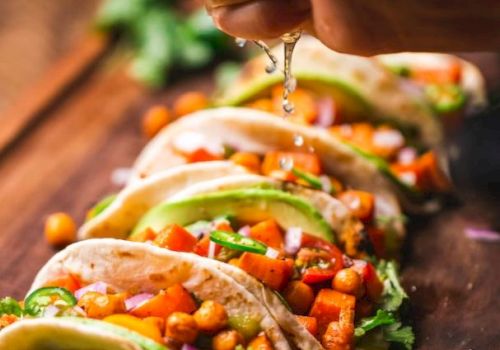 A hand is squeezing lime over a row of colorful tacos filled with chickpeas, vegetables, and herbs on a wooden board ready to be served.