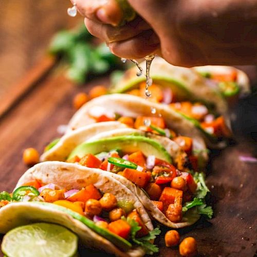 A hand is squeezing lime over a row of colorful tacos filled with chickpeas, vegetables, and herbs on a wooden board ready to be served.