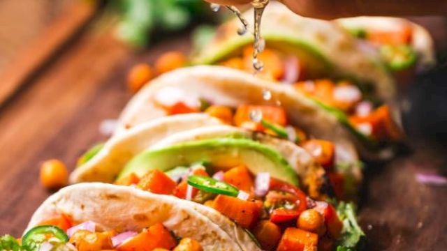 A hand is squeezing lime over a row of colorful tacos filled with chickpeas, vegetables, and herbs on a wooden board ready to be served.