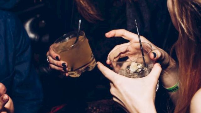 A group of people are socializing, holding drinks, and laughing together in a dimly lit environment, possibly a bar or a party setting.
