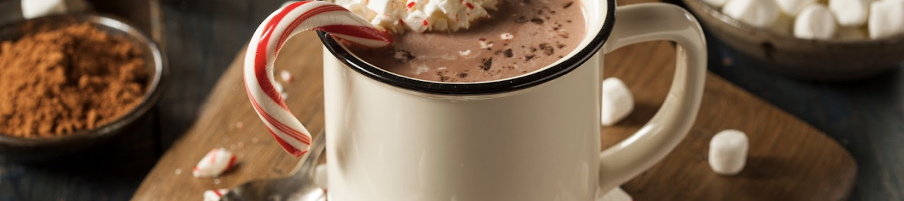 A cozy mug of hot chocolate topped with whipped cream, mini marshmallows, and a candy cane, surrounded by cocoa, peppermint sticks, and marshmallows.
