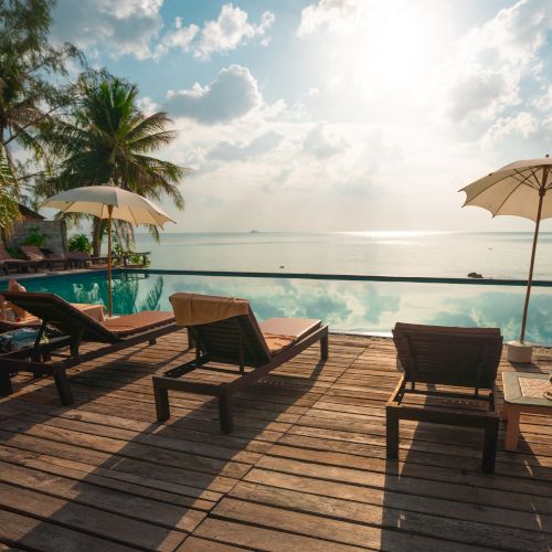 The image shows a serene poolside scene with lounge chairs, umbrellas, and palm trees overlooking a calm ocean, under a bright, partly cloudy sky.