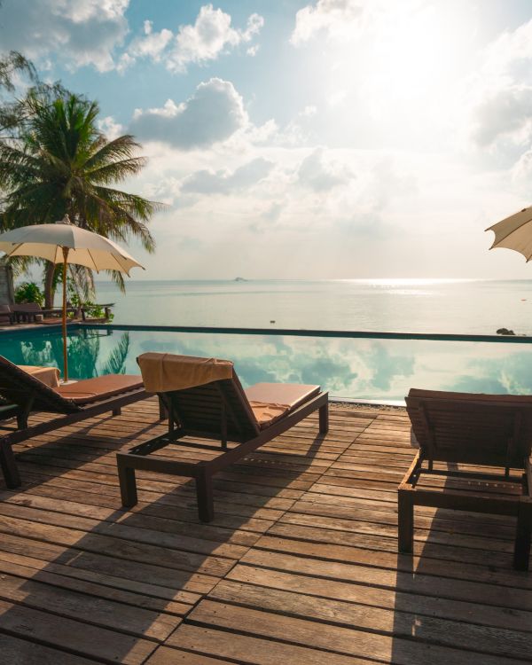The image shows a serene poolside scene with lounge chairs, umbrellas, and palm trees overlooking a calm ocean, under a bright, partly cloudy sky.