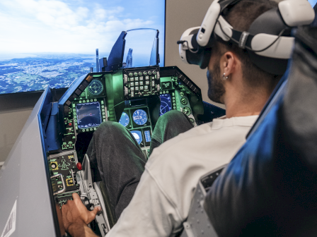A person wearing a flight-sim headset sits in a cockpit simulator, gripping the controls as dual screens show an aerial view of a sky and horizon.