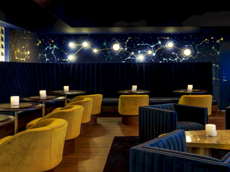 A chic lounge with blue and yellow seating, warm lighting, round tables, and a starry constellation wall mural in a modern, upscale bar.