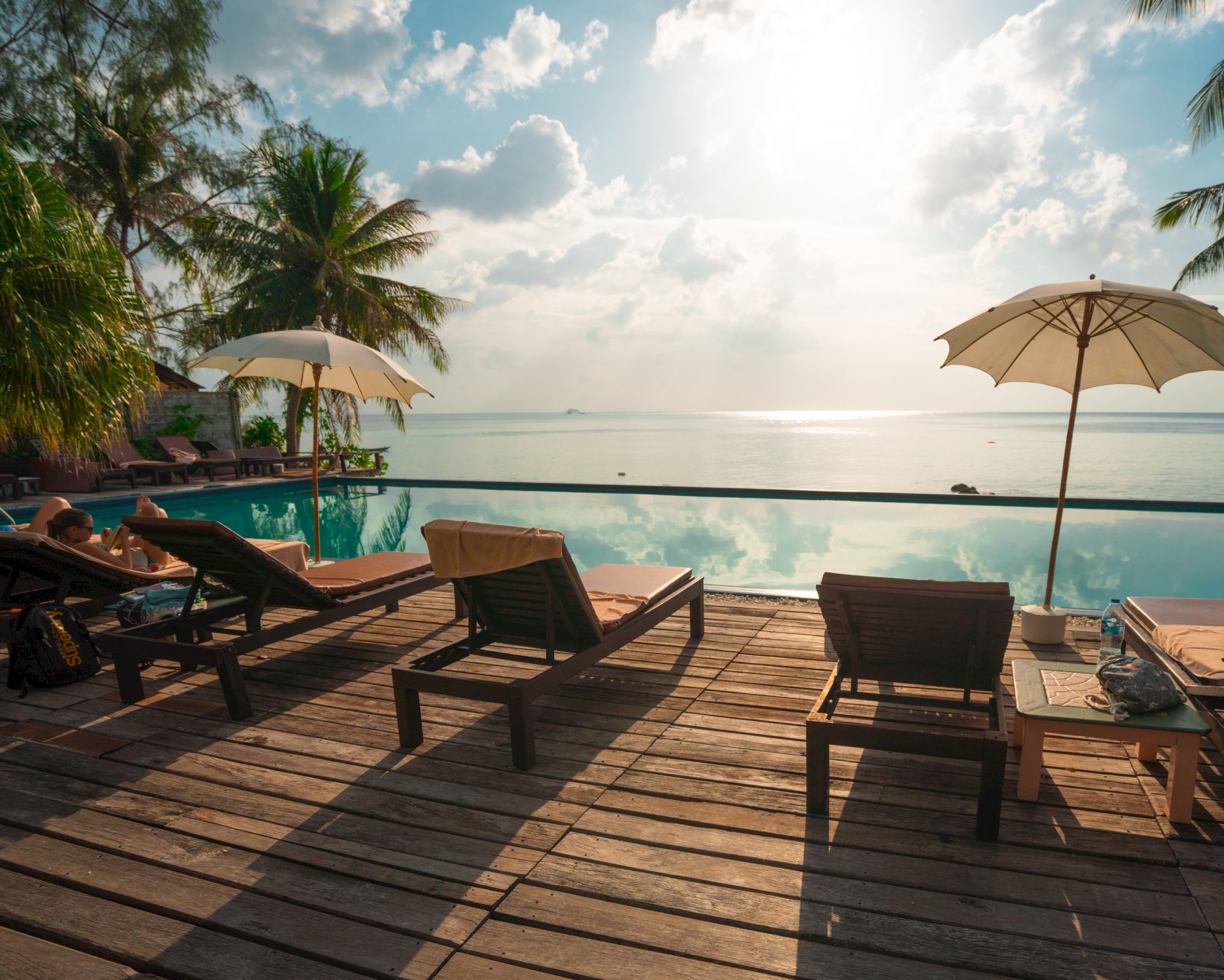 The image shows a serene poolside view with lounge chairs, umbrellas, and palm trees overlooking a calm sea, under a partly cloudy sky.