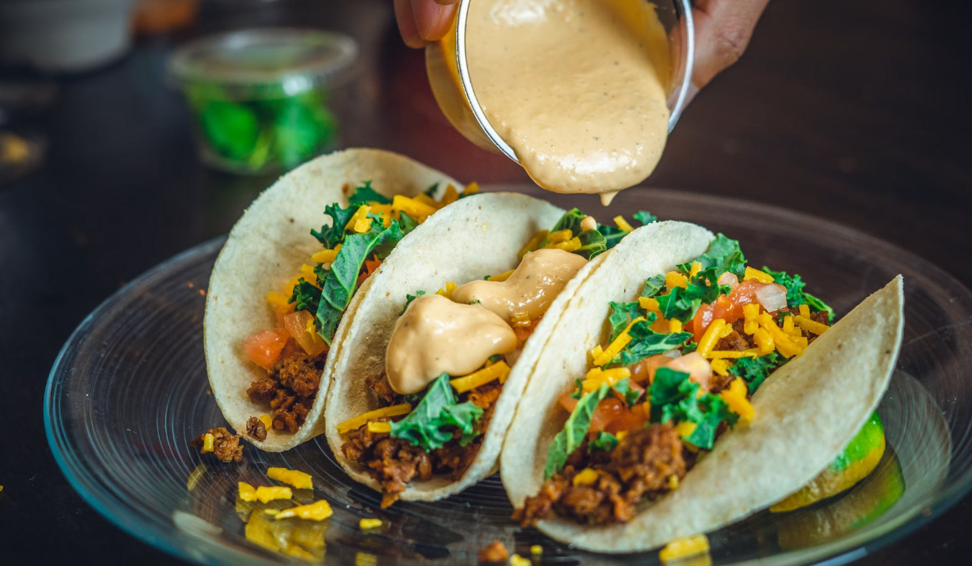 Three tacos filled with ground meat, lettuce, tomatoes, and cheese on a plate, with someone pouring sauce over the middle taco.