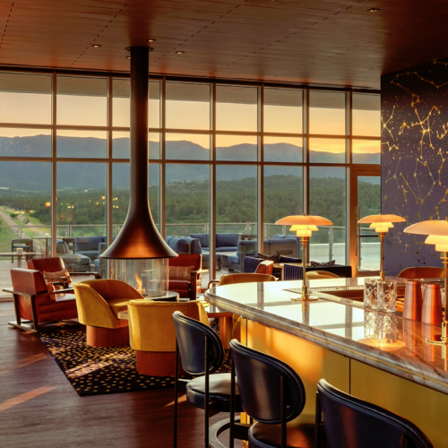 A stylish lounge with a bar, modern furniture, and a central fireplace, overlooking a scenic mountain view at sunset through large windows.