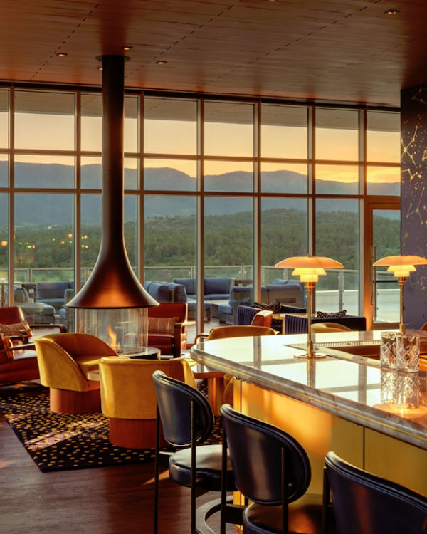 A stylish lounge with a bar, modern furniture, and a central fireplace, overlooking a scenic mountain view at sunset through large windows.