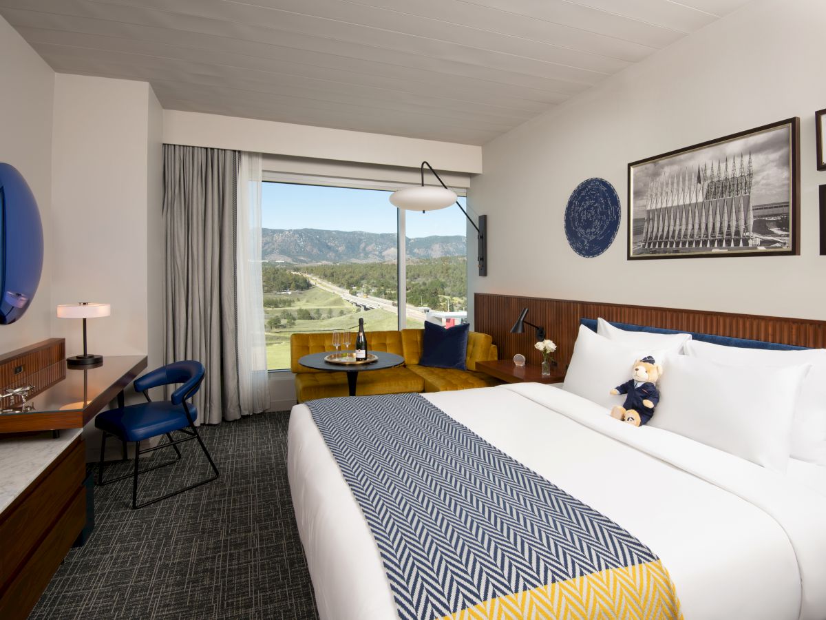 A modern hotel room with a large bed, blue and yellow accents, a scenic window view, and wall art completes this cozy space.