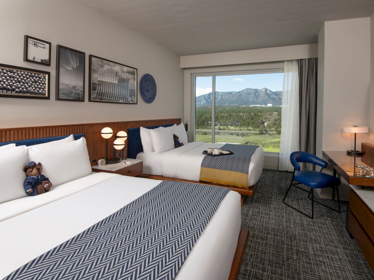 A hotel room with two beds, decorative pillows, artwork, a window view of mountains, and a small desk with a lamp.