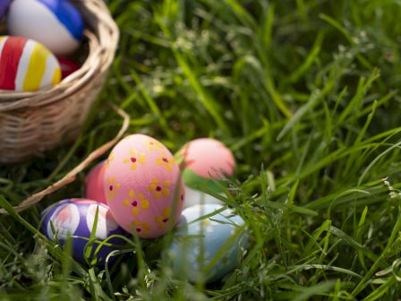 Easter eggs scattered in the grass with a wicker basket nearby, some pastel and decorated eggs resting among blades.