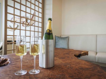 Champagne bottle in a silver cooler with two flutes on a rustic wooden table; a cozy living area with a white sofa in the background.
