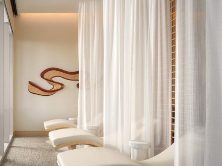 A serene spa-like room with white curtained booths and reclining lounge chairs, soft lighting, and abstract wall art in the background, calm and minimal.