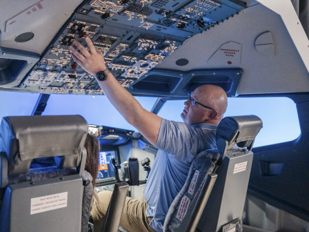 A pilot sits in the cockpit of a modern aircraft, reaching up to the overhead control panel inside the spacious cabin.