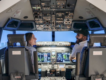 Two pilots sit and chat in the cockpit of a plane, facing each other with a bright display panel and controls between them.