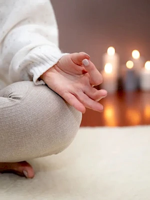A person sits cross‑legged on a soft rug, hands resting gently in their lap, with candlelight softly glowing in the background.