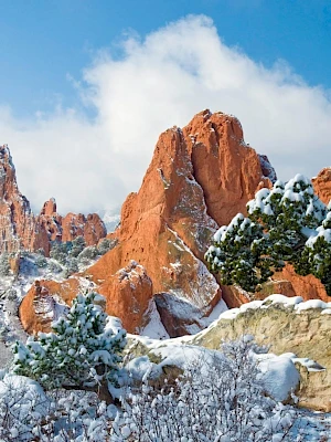 Snow-covered red rock formations with pine trees dusted in snow under a bright blue sky with scattered clouds.