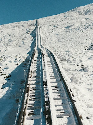 A snowy rail track climbs a hill, slick with ice and framed by white, barren slopes under a clear blue sky.