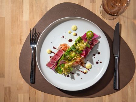 A beautifully plated dish with colorful ingredients, sauce drizzles, garnished with greens and sliced jalapeños, served with cutlery and a glass.