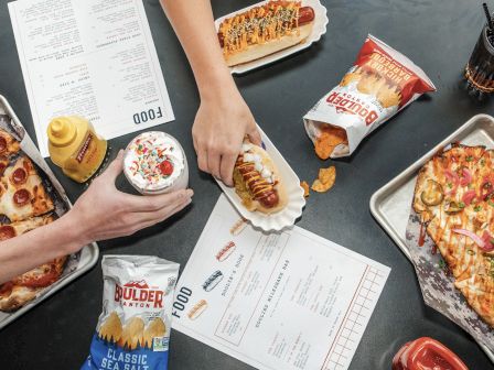 The image shows a table with pizzas, hot dogs, chips, a milkshake, soda, and menus. Hands are reaching for food items.