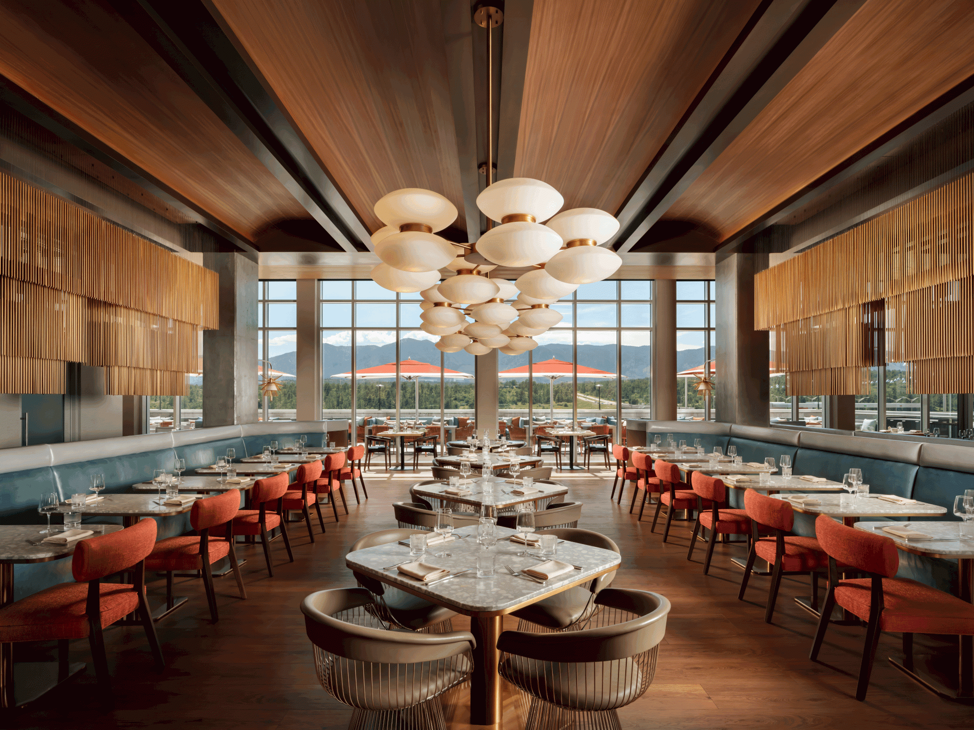A modern restaurant with elegant lighting, wooden accents, and mountain views, featuring red chairs and white table settings.