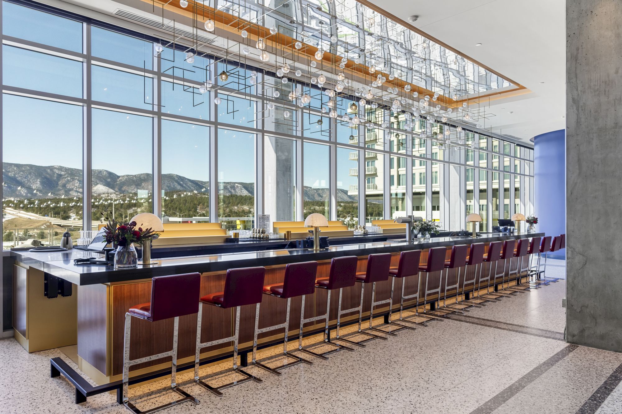 The image shows a sleek bar with red chairs, modern lighting, and large windows offering a mountain view in a contemporary setting.