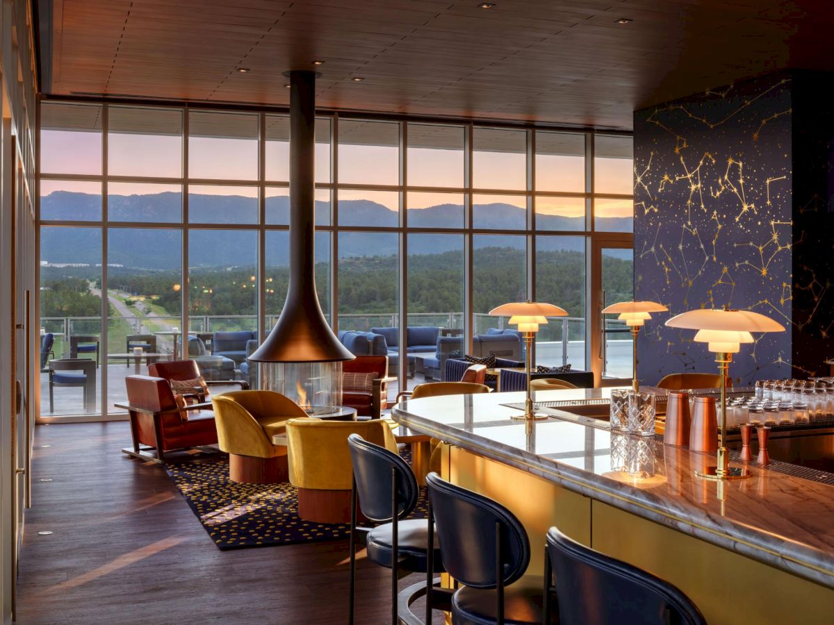 A stylish lounge with a long bar, colorful chairs, a hanging fireplace, large windows, and a scenic mountain view in the distance.